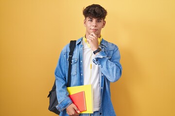 Hispanic teenager wearing student backpack and holding books with hand on chin thinking about...