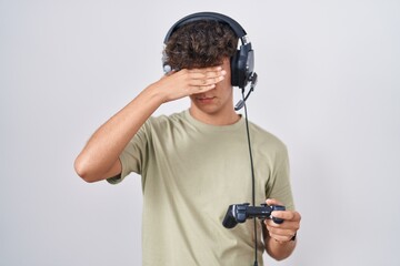 Hispanic teenager playing video game holding controller covering eyes with hand, looking serious and sad. sightless, hiding and rejection concept