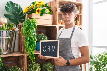 Hispanic teenager working at florist holding open sign thinking attitude and sober expression...
