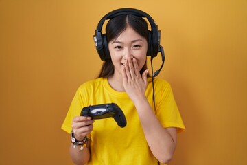 Chinese young woman playing video game holding controller laughing and embarrassed giggle covering mouth with hands, gossip and scandal concept