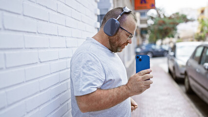 Cheerful, middle-aged, bearded caucasian man, vibing on urban street, joyfully dancing while listening to music on smartphone, letting the rhythm flow through his headphones.