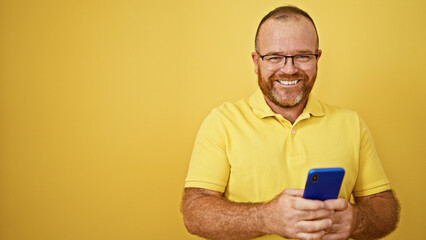 Cheerful bearded caucasian man with glasses, joyfully using smartphone, typing, sending happy texts over yellow isolated background