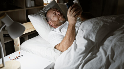 Handsome, middle-aged, bearded caucasian man relaxing in bed, using his smartphone in the warm glow...
