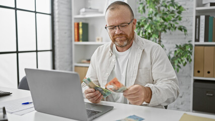 Confident, smiling caucasian man counting australian dollars at his office desk using laptop, a...