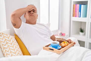 Young hispanic man eating breakfast in the bed stressed and frustrated with hand on head, surprised and angry face