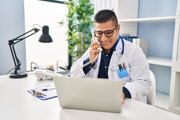 Young latin man doctor using laptop talking on telephone at clinic - obrazy, fototapety, plakaty