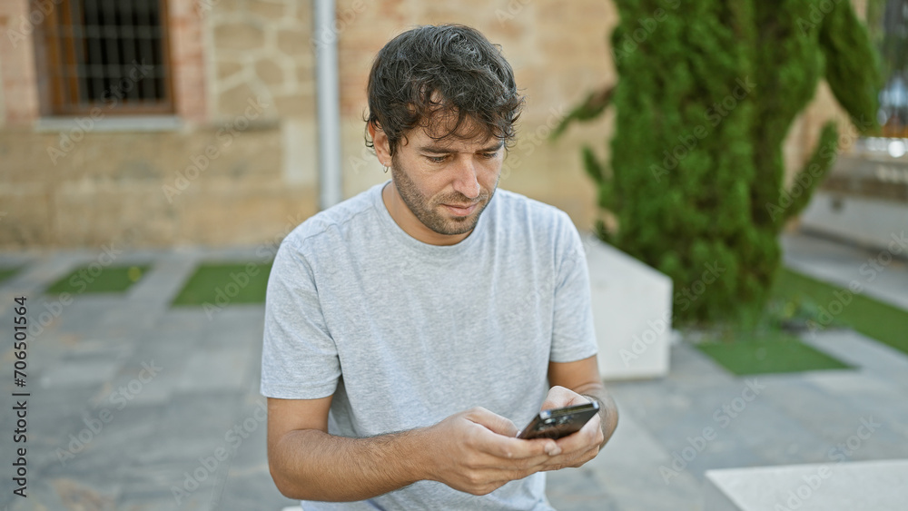 Canvas Prints Handsome, young hispanic guy with blond beard gets lost in his smartphone screen while sitting and texting in the lush city park, a portrait of concentrated technology use amidst the green outdoors