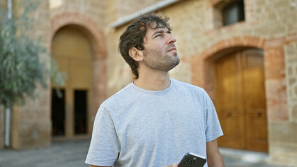 Relaxed, bearded young man, engrossed in his smartphone, gazes at the city sky in a green park - a...