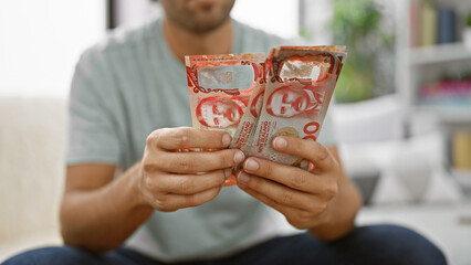 Young hispanic man engrossed in counting stacks of new zealand dollars on his living room sofa,...