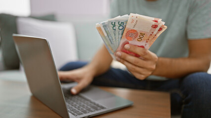 Young man, sitting on his sofa at home, handling yuan banknotes while engrossed in online finance...