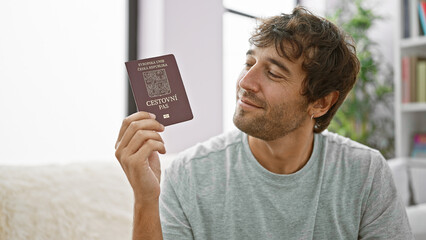 Confident bearded young man sitting on his living room sofa, grinning wide with czech republic passport, overflowing with happiness, ready to embark on an exciting vacation - obrazy, fototapety, plakaty