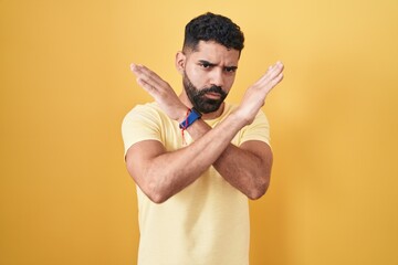 Hispanic man with beard standing over yellow background rejection expression crossing arms doing negative sign, angry face