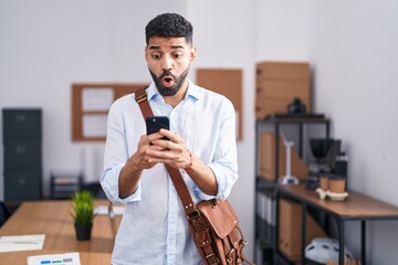 Hispanic man with beard using smartphone at the office afraid and shocked with surprise and amazed...