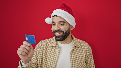 Young hispanic man wearing christmas hat holding credit card over isolated red background