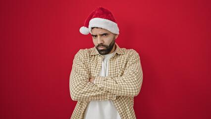 Young hispanic man wearing christmas hat standing with arms crossed gesture and unhappy expression over isolated red background