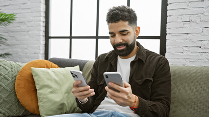 A young hispanic man with a beard smiles while using two smartphones in a modern living room, implying connectivity and multitasking.