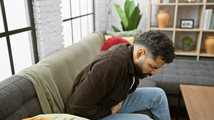 Young hispanic man with beard feeling stomachache, sitting on couch in home living room interior.