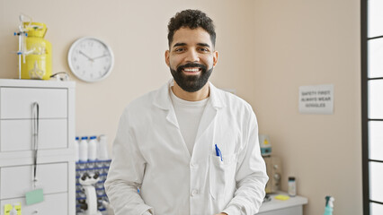 Handsome hispanic man with beard wearing a lab coat standing in a bright hospital room - obrazy, fototapety, plakaty