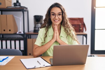 Young hispanic woman working at the office wearing glasses praying with hands together asking for forgiveness smiling confident.