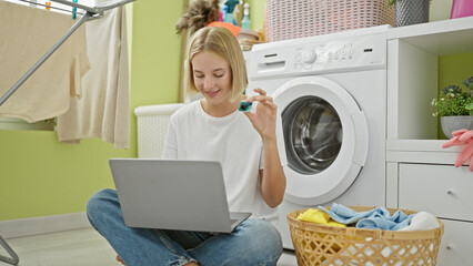 Young blonde woman using laptop holding detergent bag at laundry room