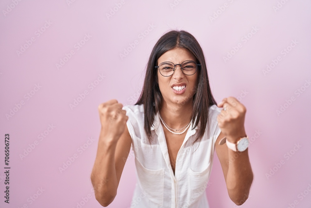 Sticker Brunette young woman standing over pink background wearing glasses angry and mad raising fists frustrated and furious while shouting with anger. rage and aggressive concept.