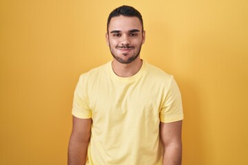 Young hispanic man standing over yellow background puffing cheeks with funny face. mouth inflated with air, crazy expression. - obrazy, fototapety, plakaty