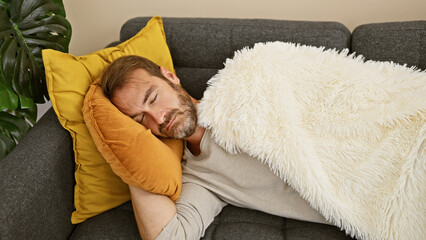 A middle-aged man naps comfortably on a couch with a white blanket and yellow pillows, highlighting relaxation at home.