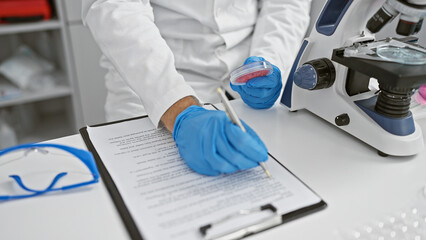 Cropped view of a scientist in a laboratory analyzing specimens with a microscope and taking notes.