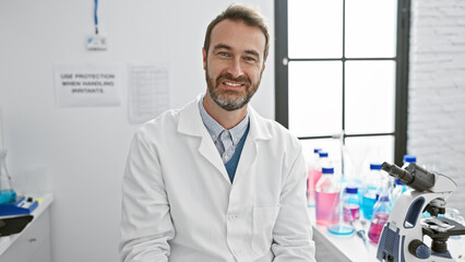 Smiling middle-aged hispanic man in lab coat indoors with microscope and lab equipment, portraying...