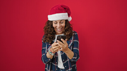 Middle age hispanic woman wearing christmas hat using smartphone over isolated red background