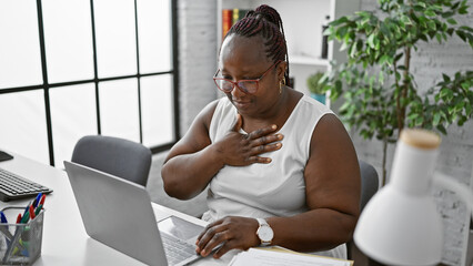 Smiling african american woman confidently working online in the office, equipped with her laptop, hand on chest, embodying success in business - obrazy, fototapety, plakaty