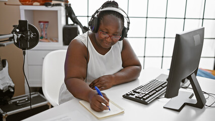 Smiling african american woman, a confident musician, composing a soulful song in a music studio,...