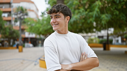 Cheerful young hispanic teenager confidently standing in city park, arms crossed, happily looking...