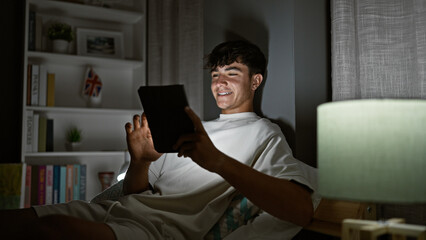Young hispanic teenager smiling while comfortably sitting on his bed, enjoying the morning in his...