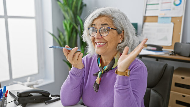 A smiling mature woman, holding a smartphone, in a bright office setting