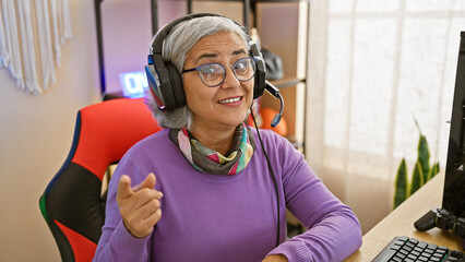 A smiling mature woman with grey hair wearing a headset in a gaming room with a colorful chair and...