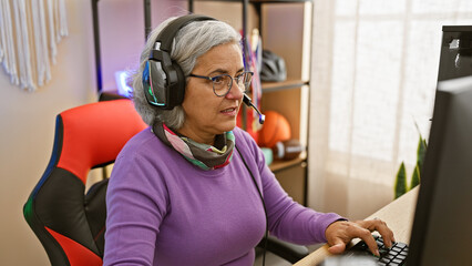 A middle-aged woman engages in gaming indoors, wearing headphones and glasses, illuminating...