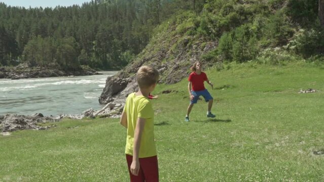 Cute little children playing with frisbee outdoors on sunny day. Against the backdrop of mountains, on the coast of a mountain river. Slow motion. Young Boys throwing a red frisbee disk. 