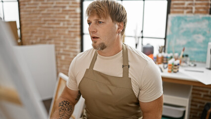 Blond man with beard in apron focused on painting in art studio interior.