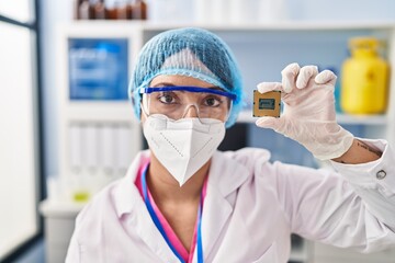 Fototapeta na wymiar Young beautiful hispanic woman scientist wearing medical mask holding cpu processor chip at pharmacy
