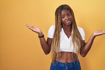 African american woman with braided hair standing over yellow background clueless and confused...