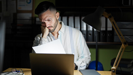 Young hispanic man business worker using laptop reading document looking upset at the office