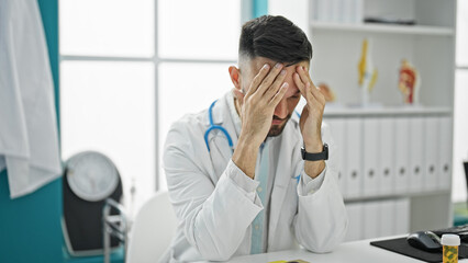 Young hispanic man doctor sitting on table stressed at the clinic