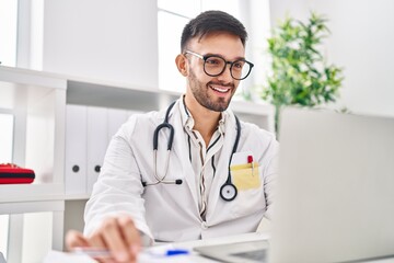 Young hispanic man doctor smiling confident using laptop at clinic