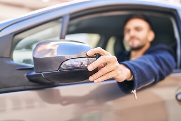 Young hispanic man touching rearview sitting on car at street