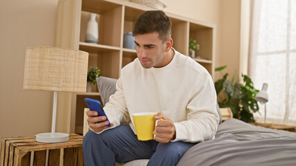 Charming young hispanic man immersed in texting on smartphone, comfortably relaxed in bedroom,...
