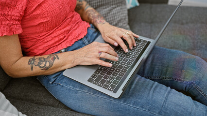 Naklejka premium At home, a hardworking woman's hands click away on her laptop, comfortably ensconced on her cozy sofa amidst the familiar backdrop of her living room, fully utilising technology to work online.