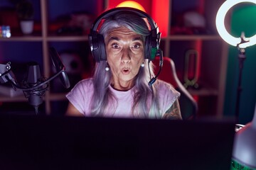 Middle age woman with tattoos playing video games wearing headphones scared and amazed with open...