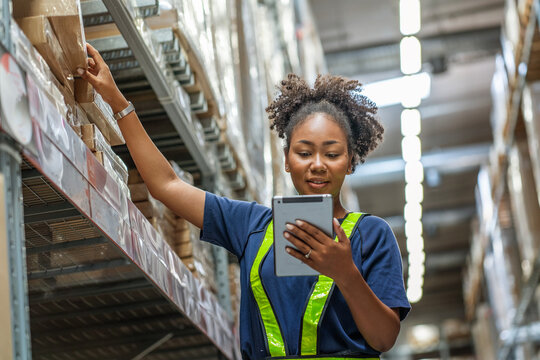 Black African woman employee holding tablet walks through inventory and checks orders from customers, picks up boxes from shelves to deliver documents to customers in warehouse wholesale store.