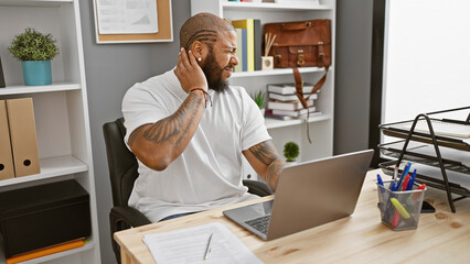 African american man with beard feeling neck pain at office workstation with laptop and decorative plant in background - obrazy, fototapety, plakaty